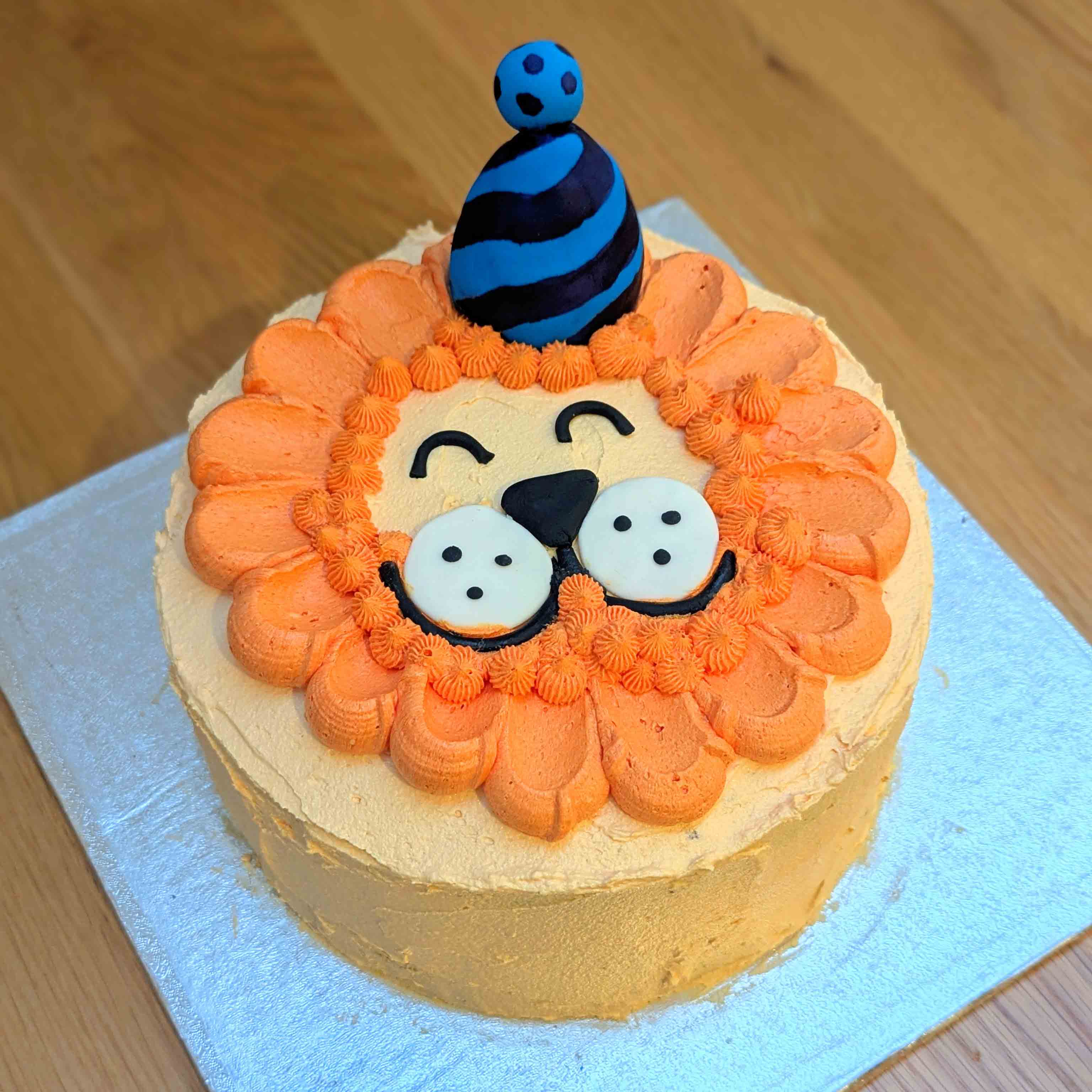 Smiling lion face cake with an orange piped mane and a striped party hat topper